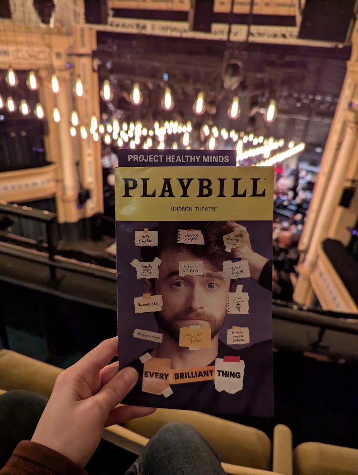 Holding my playbill for Every Brilliant Thing starring Daniel Radclife partially showing my view of the stage from the second row balcony seat. They had a lot of oversized light bulbs with the visible filament hanging from the ceiling as decoration.