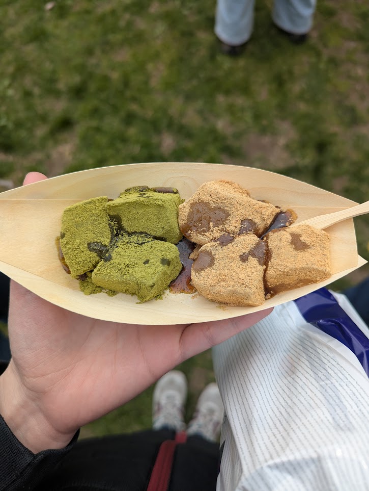 Six pieces of mochi in a bamboo boat. Three are covered in green powder and three are covered in brown powder.