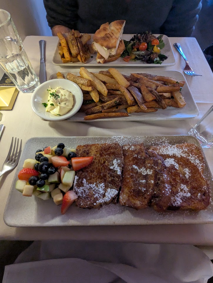 Plate of 3 slices of French toast covered in powdered sugar along with assorted fruit. There's a plate in the middle just slammed with more wedge potato fries. In the back my husband has a plate with a panini, inexplicably more fries, and a house salad.